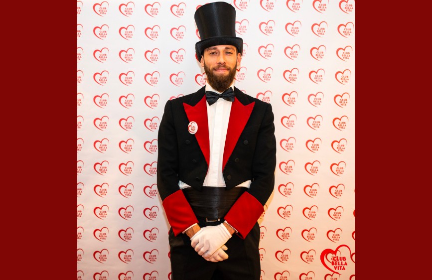 Nella foto è ritratto un uomo in posa frontale, vestito con un elegante frac nero con ampi risvolti rossi, guanti bianchi, papillon e un cilindro nero in testa. Indossa un distintivo con la scritta "Club della Vita" su sfondo bianco e rosso. Alle sue spalle, il fondale è completamente ricoperto da un motivo ripetuto di cuori rossi con la stessa scritta. L'inquadratura è centrata e l’illuminazione uniforme, adatta a un contesto formale o celebrativo.