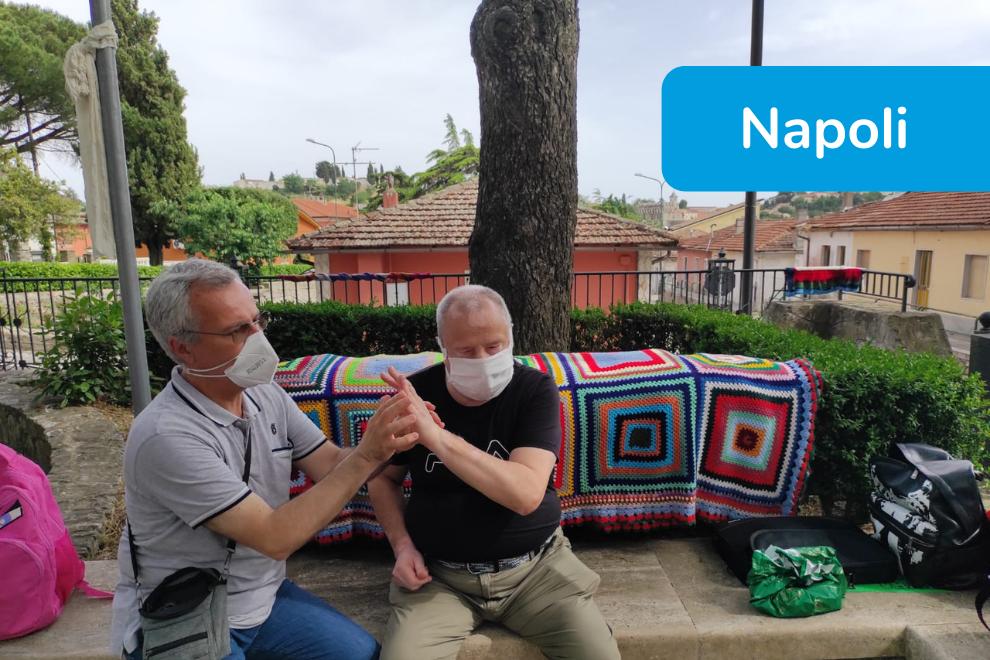 Yarn Bombing - Napoli - un utente adulto siede su una panchina dove sono esposti i manufatti colorati e un operatore comunica con lui in Malossi indossando la mascherina