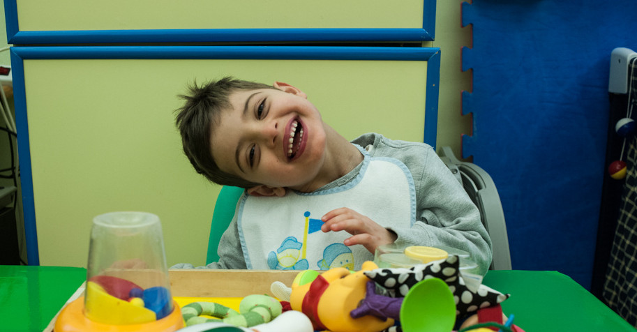 Foto di Benito da bambino (6 anni circa) alla Lega del FIlo d'Oro: piega la testa da un lato e fa un bel sorriso seduto davanti a un vassoio pieno di giocattoli colorati
