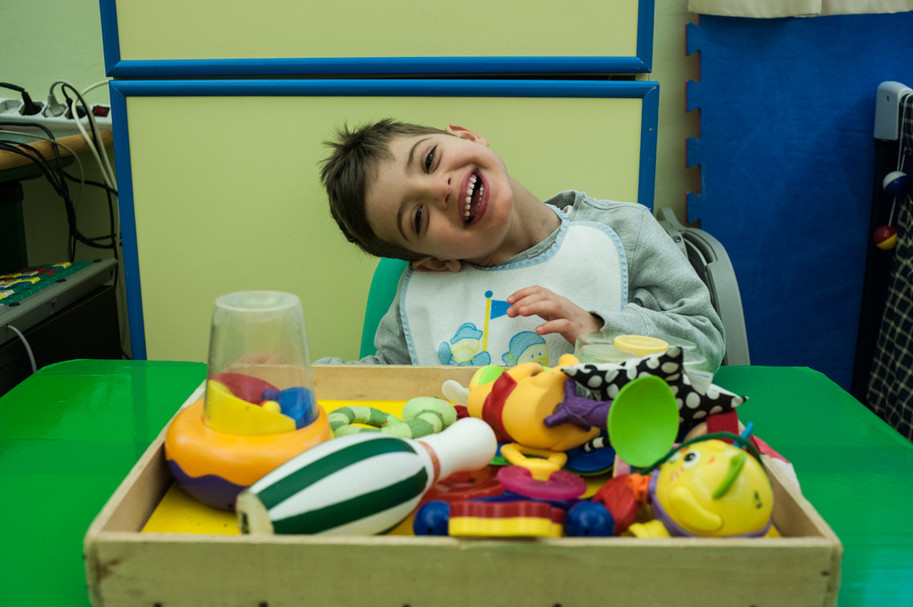 Foto di Benito da bambino (6 anni circa) alla Lega del FIlo d'Oro: piega la testa da un lato e fa un bel sorriso seduto davanti a un vassoio pieno di giocattoli colorati
