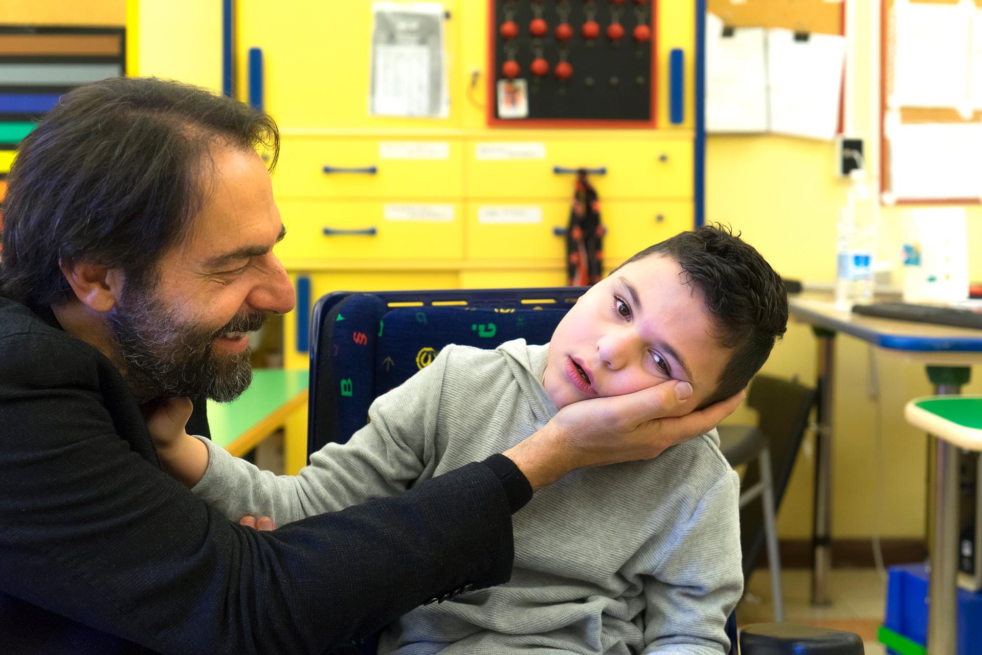Neri Marcorè sorridente dà una carezza a un bambino sordocieco, che adagia il volto sulla sua mano.