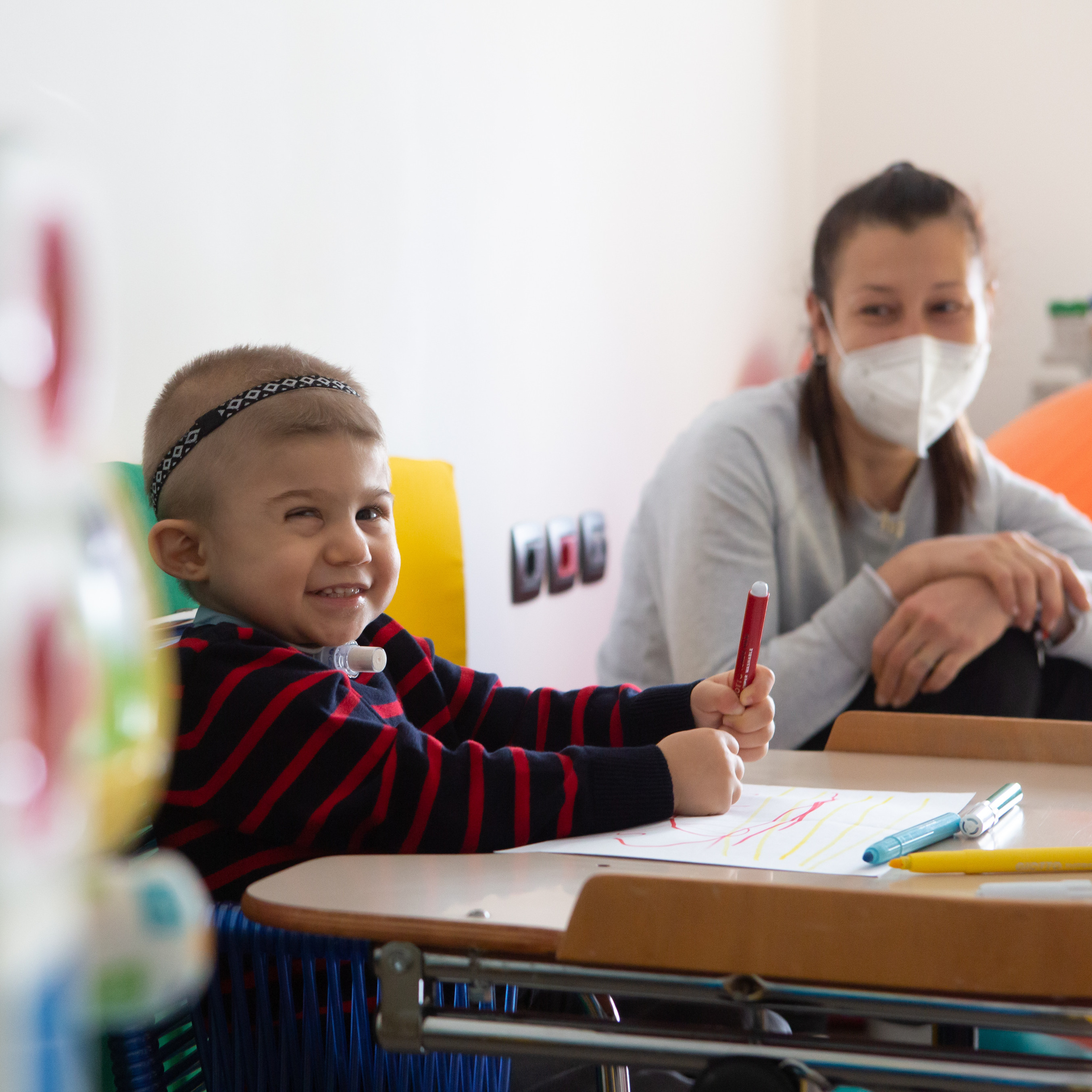 Il piccolo Berk ospite del Centro Diagnostico fa un sorriso mentre disegna con i pennarelli su un foglio. La sua mamma, con indosso una mascherina anti covid, lo guarda con amore seduta di fianco a lui