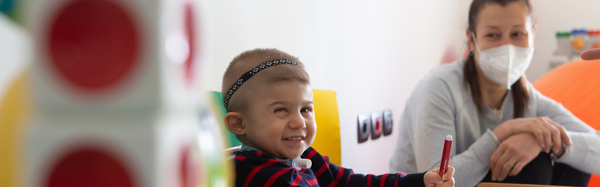 Il piccolo Berk ospite del Centro Diagnostico fa un sorriso mentre disegna con i pennarelli su un foglio. La sua mamma, con indosso una mascherina anti covid, lo guarda con amore seduta di fianco a lui