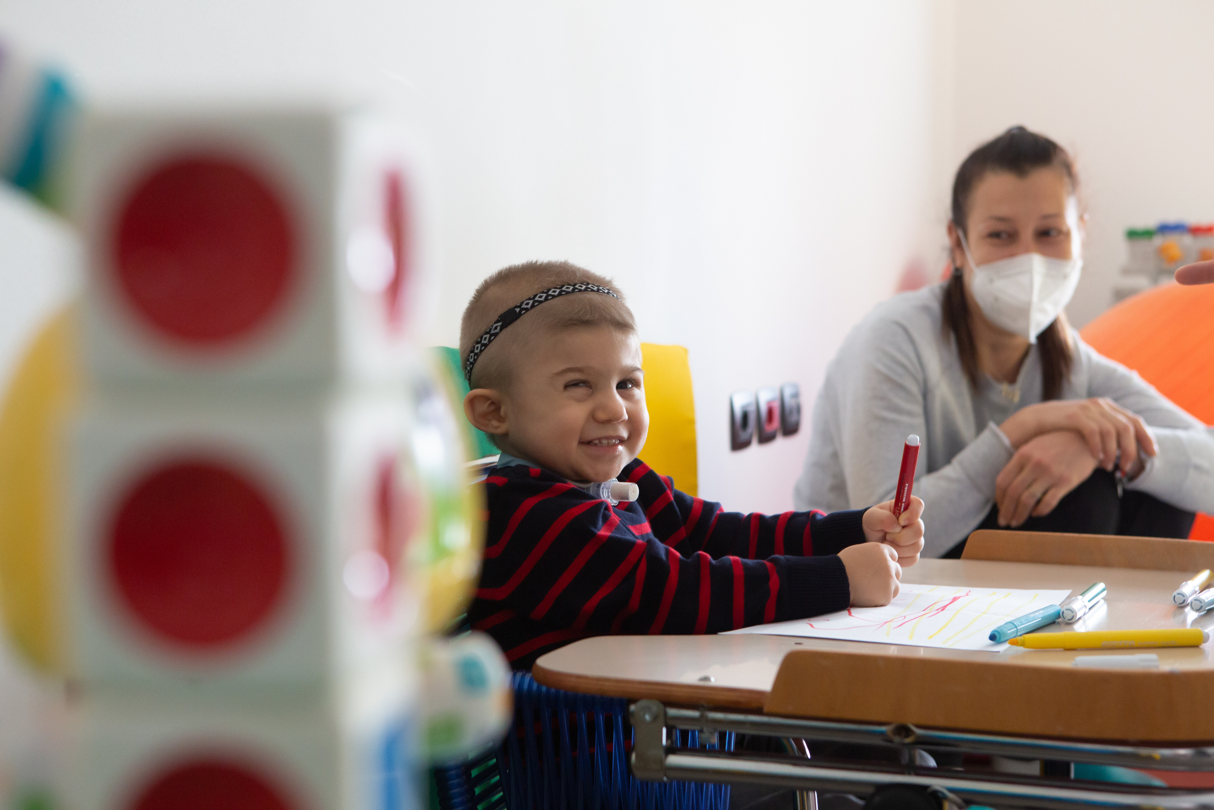 Il piccolo Berk ospite del Centro Diagnostico fa un sorriso mentre disegna con i pennarelli su un foglio. La sua mamma, con indosso una mascherina anti covid, lo guarda con amore seduta di fianco a lui