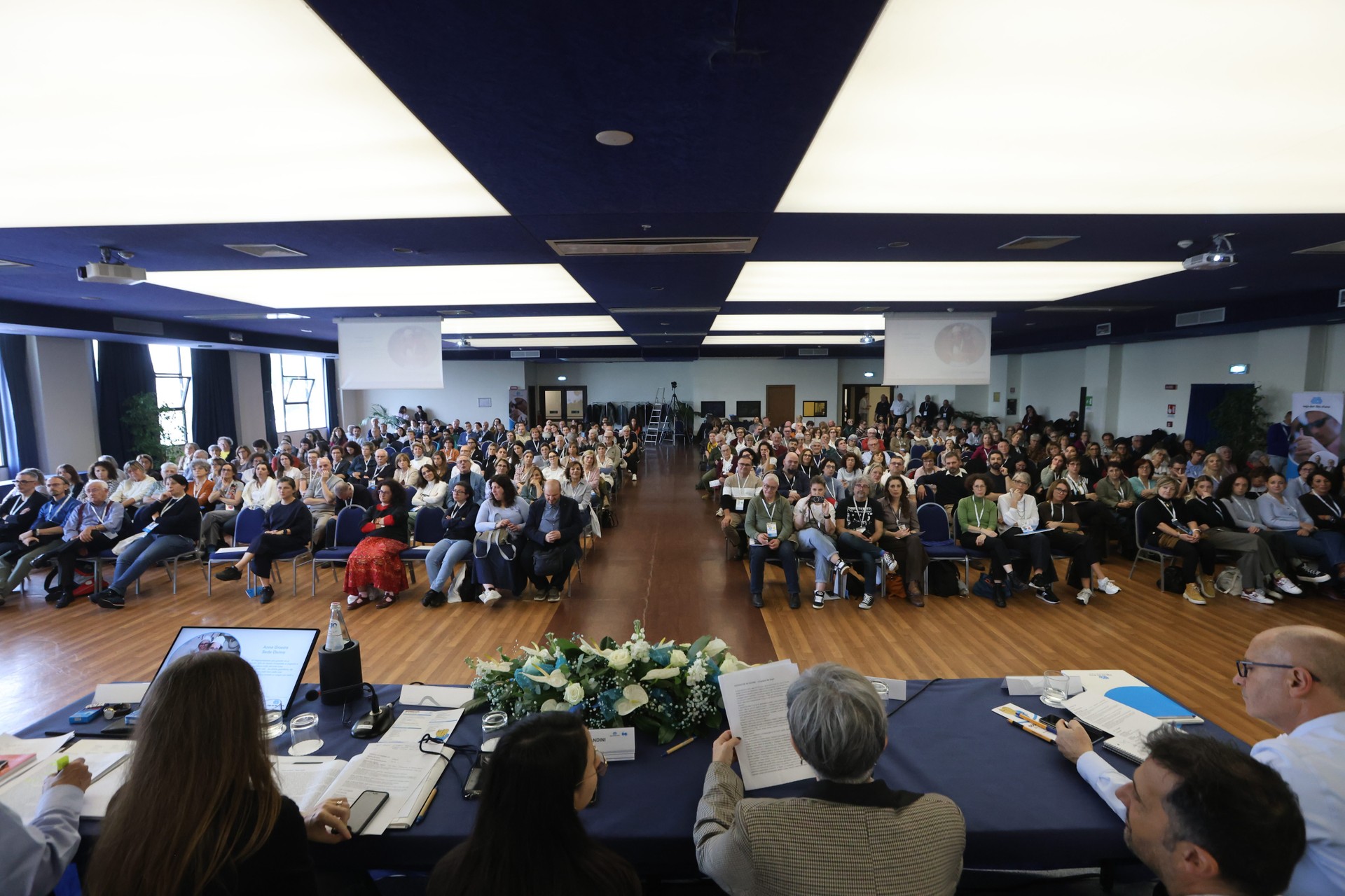 Fotografia scattata durante le testimonianze al Forum dei Volontari. La platea è seduta ai propri posti mentre il punto di vista, dal tavolo dei relatori, restituisce un momento di ascolto, condivisione e partecipazione.