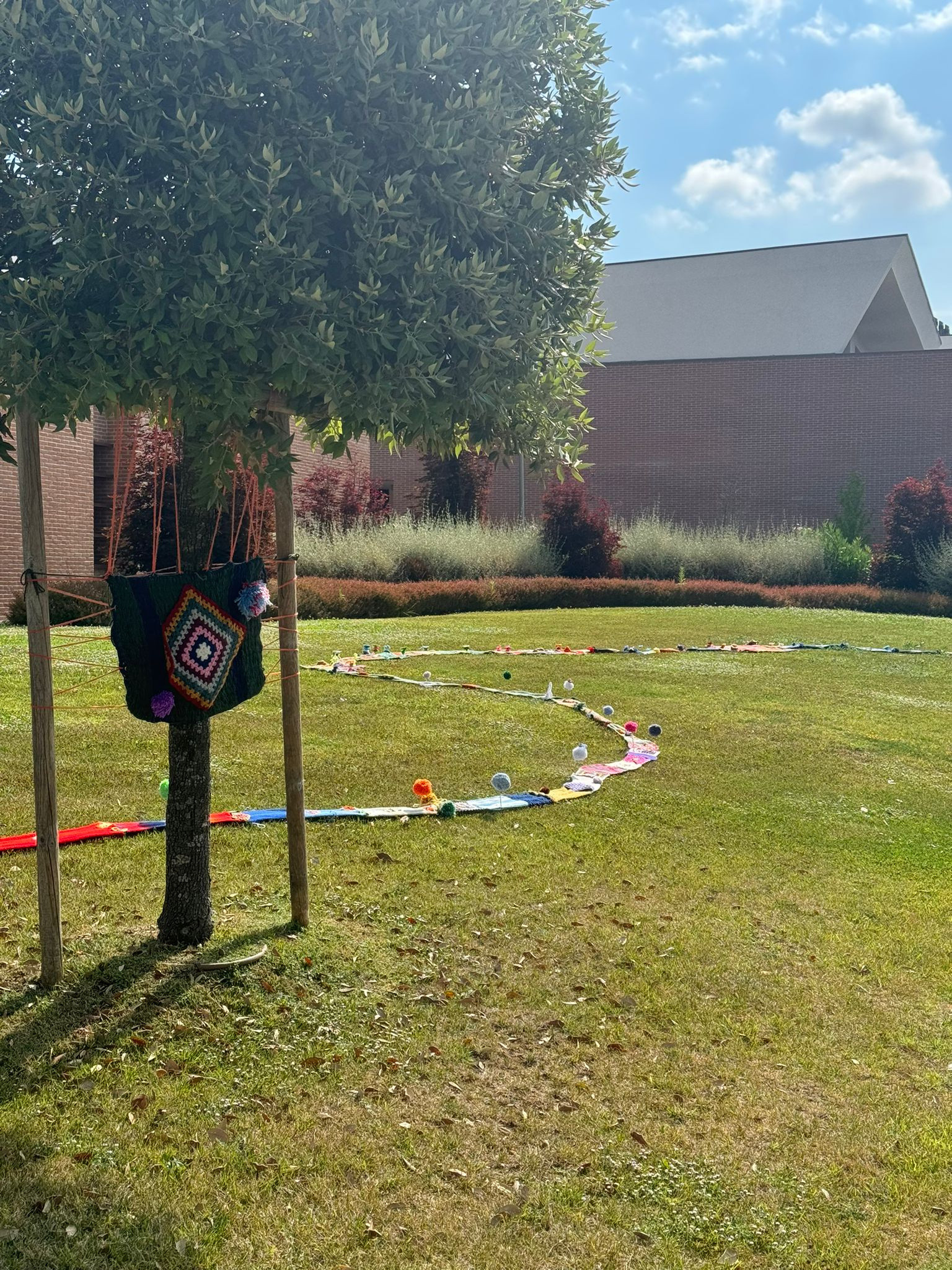 Su un prato illuminato dal sole si snoda una lunga serpentina fatta a maglia. A un albero vicino è appesa una borsa nera, anch’essa realizzata a maglia, con un vivace ricamo colorato al centro. 