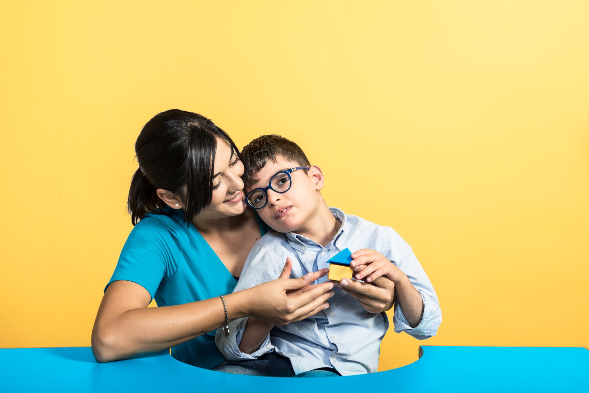 Immagine della campagna "Un contatto che vale": il piccolo Benny, utente del Centro di Osimo, tiene in mano una casetta di legno gialla e blu, seduto in braccio alla sua operatrice che gli sorride