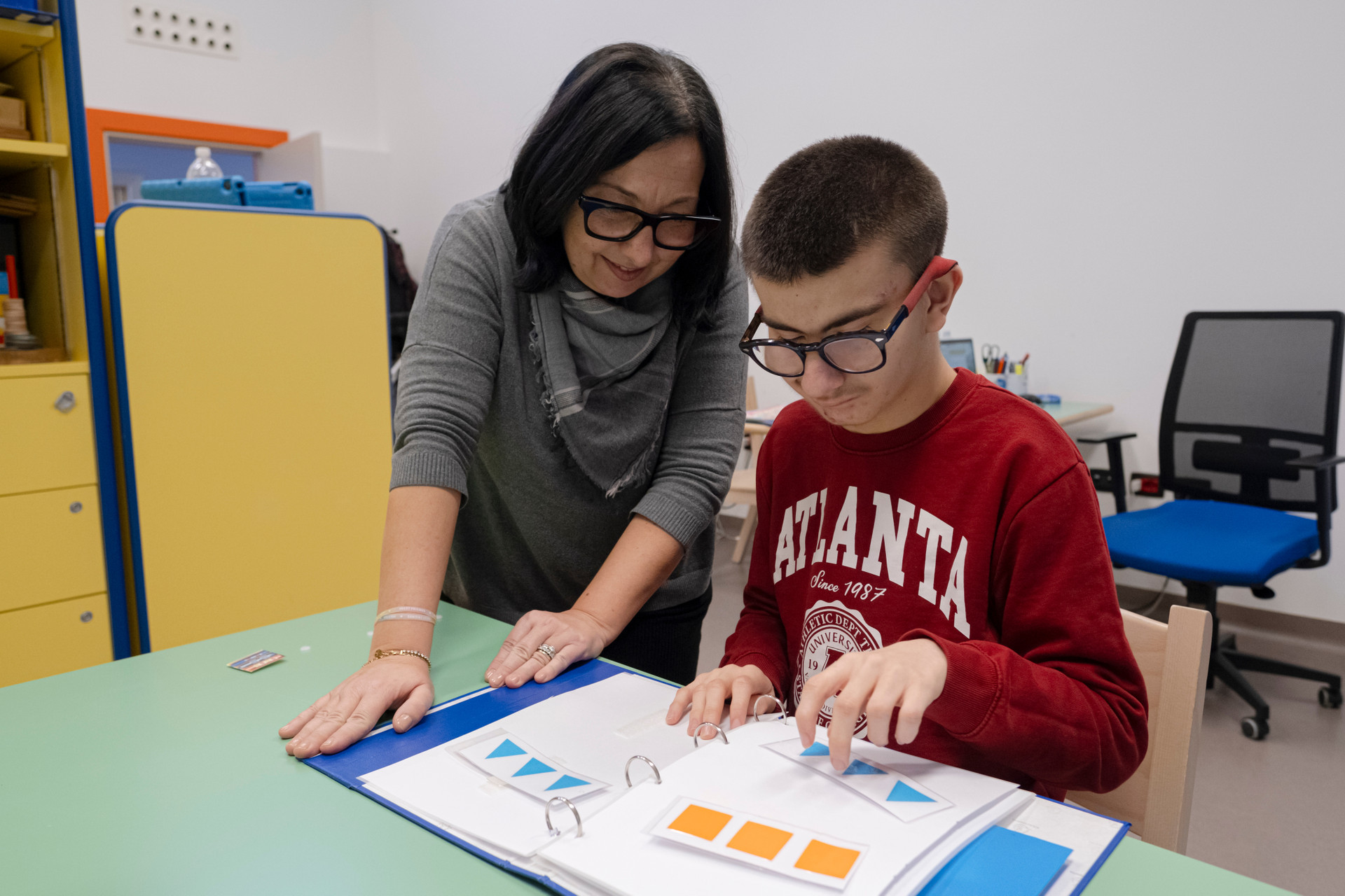 Biagio Luigi, durante l'ultimo Trattamento Intensivo, lo scorso Gennaio 2025. Qui è in aula con la sua educatrice mentre fanno attività: Luigi associa le figura uguali nel suo quadernone blu delle attività. L'educatrice lo supporta.