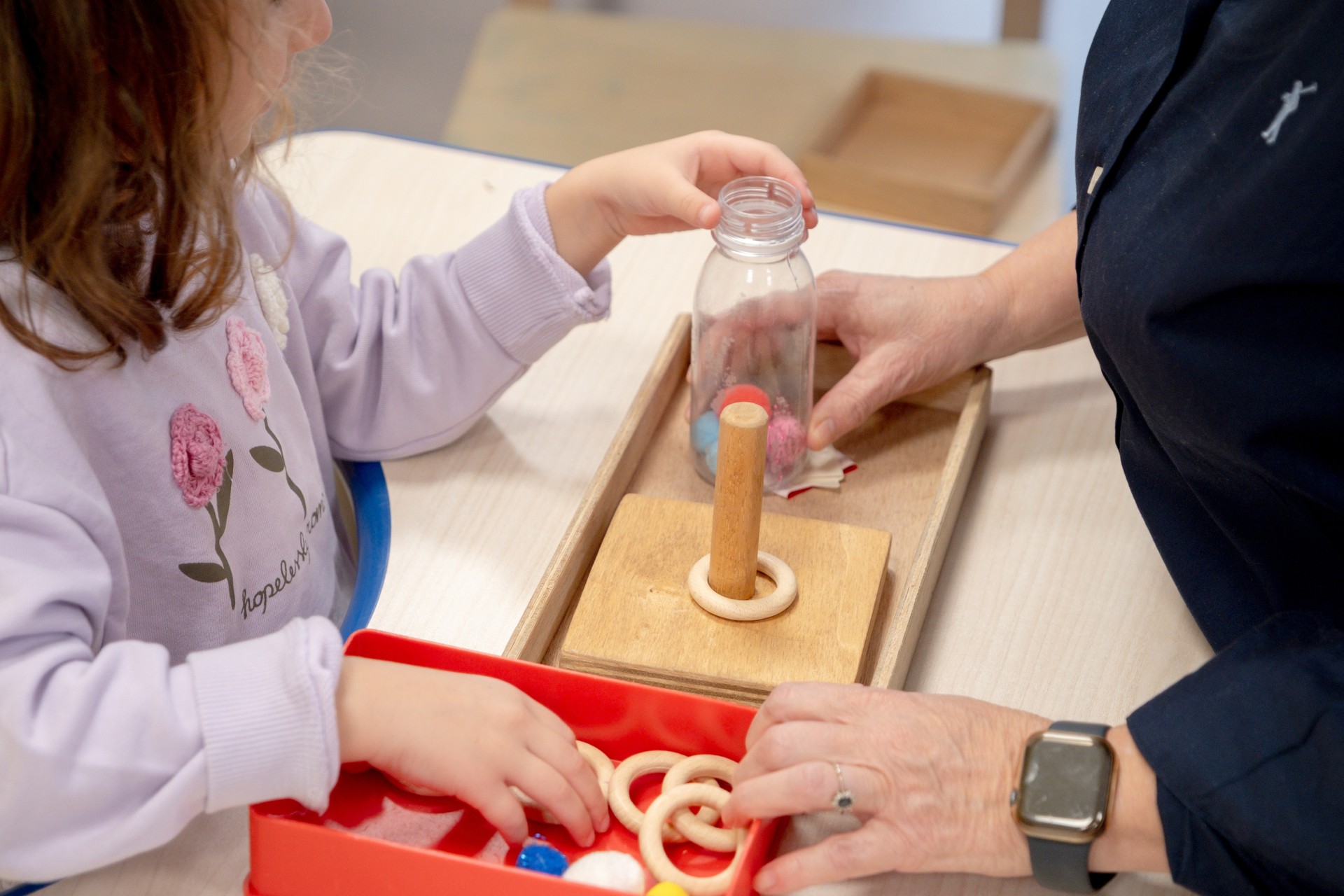 Una bambina ritratta durante un’attività educativa, è seduta al tavolo insieme a una educatrice della Lega del Filo d’Oro. Utilizza anelli di legno, una base a incastro e altri materiali contenuti in una vaschetta e in una bottiglia trasparente.
