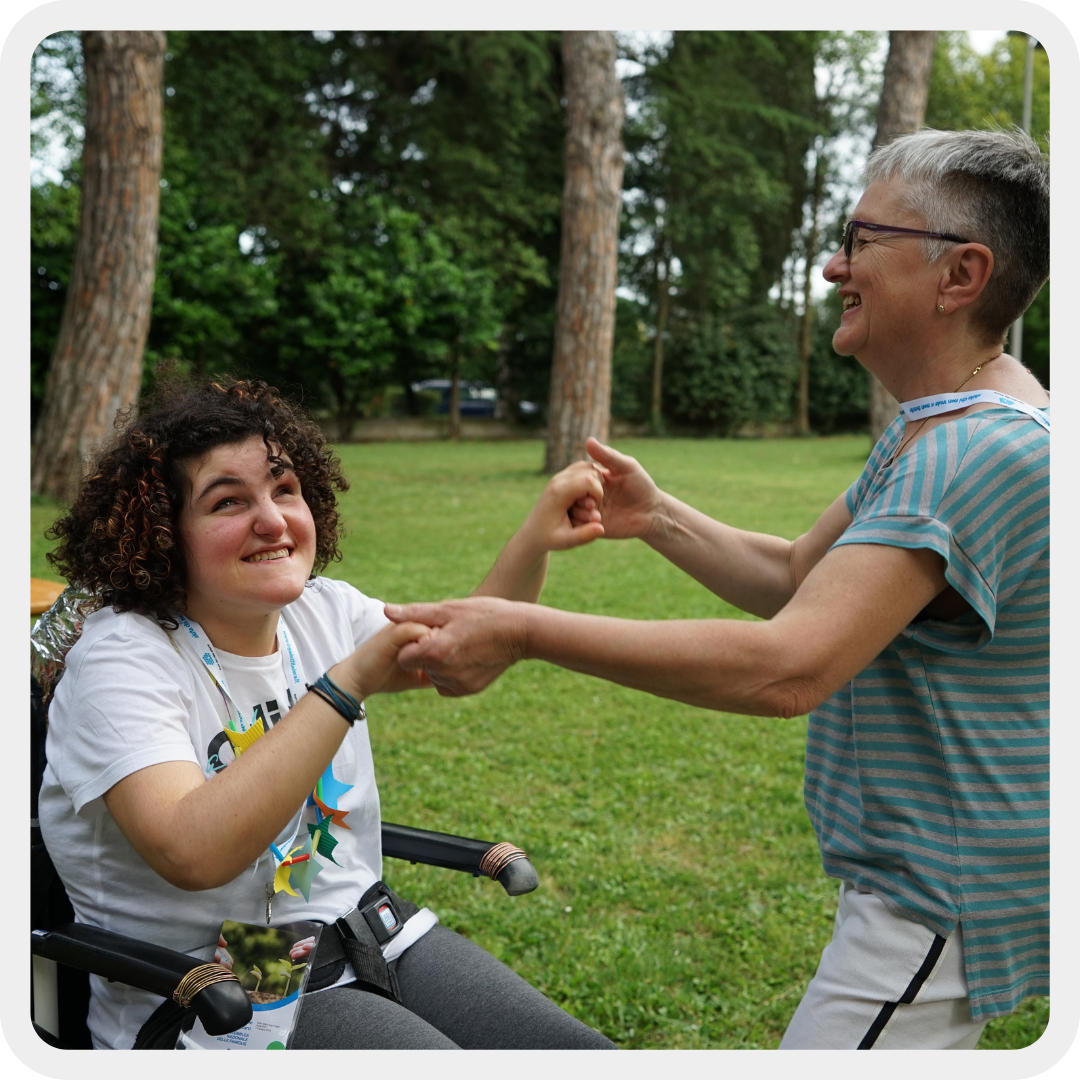 Sara, una giovane ragazza sordocieca con disabilità motorie, si diverte in un giardino con una volontaria. Si tengono per mano e sorridono entrambe.