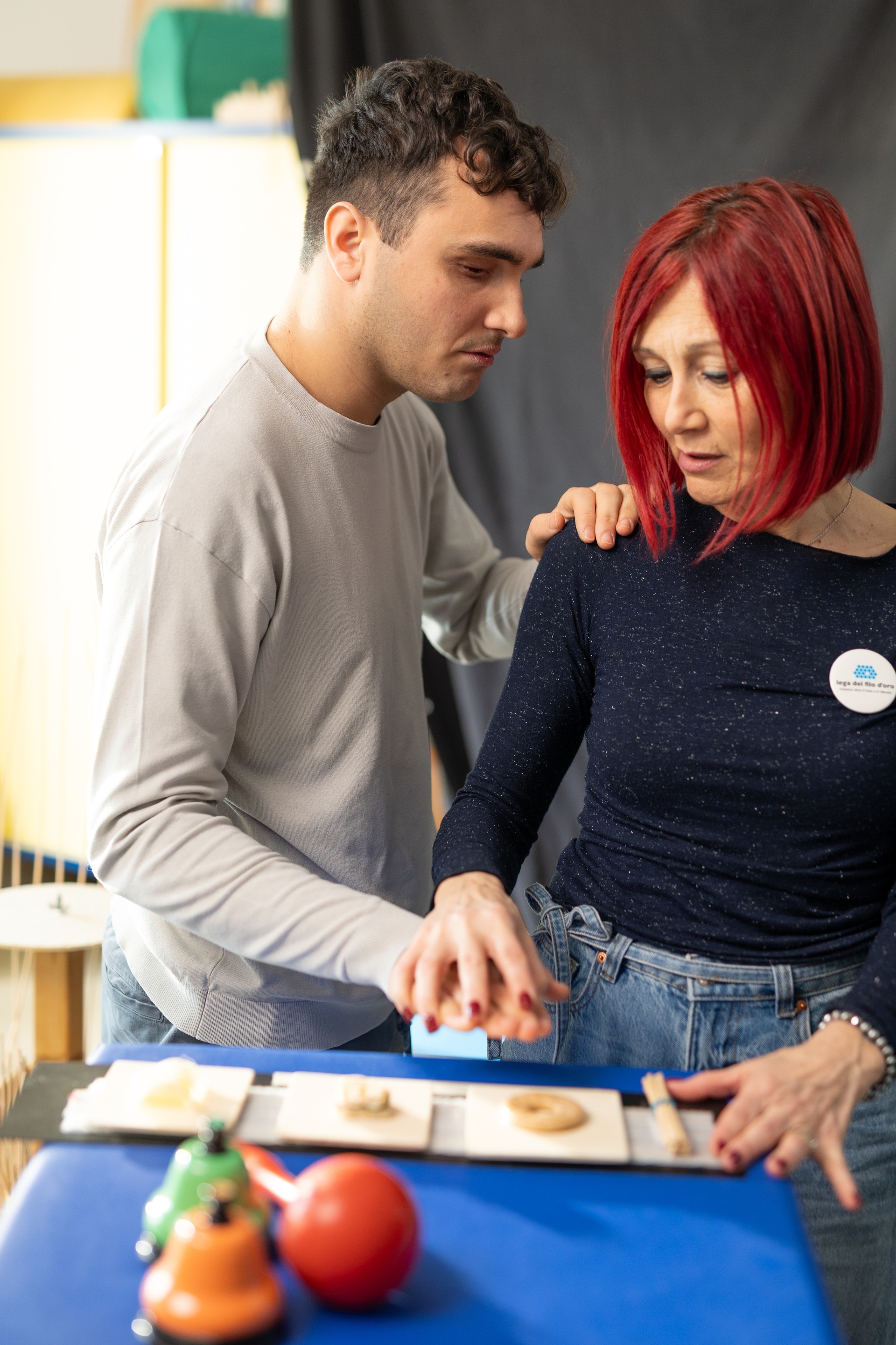 Francesco è in piedi a fiano della sua educatrice. Lei lo tiene per mano e lo aiuta ad allenare il suo senso tattile con un libro