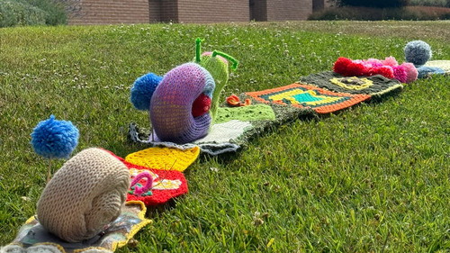Su un prato verde si snoda una lunga serpentina colorata fatta a maglia. Sulla sua superficie sono posizionate lumachine realizzate all’uncinetto e pompon colorati.