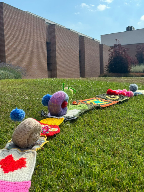 Su un prato verde si snoda una lunga serpentina colorata fatta a maglia. Sulla sua superficie sono posizionate lumachine realizzate all’uncinetto e pompon colorati. 