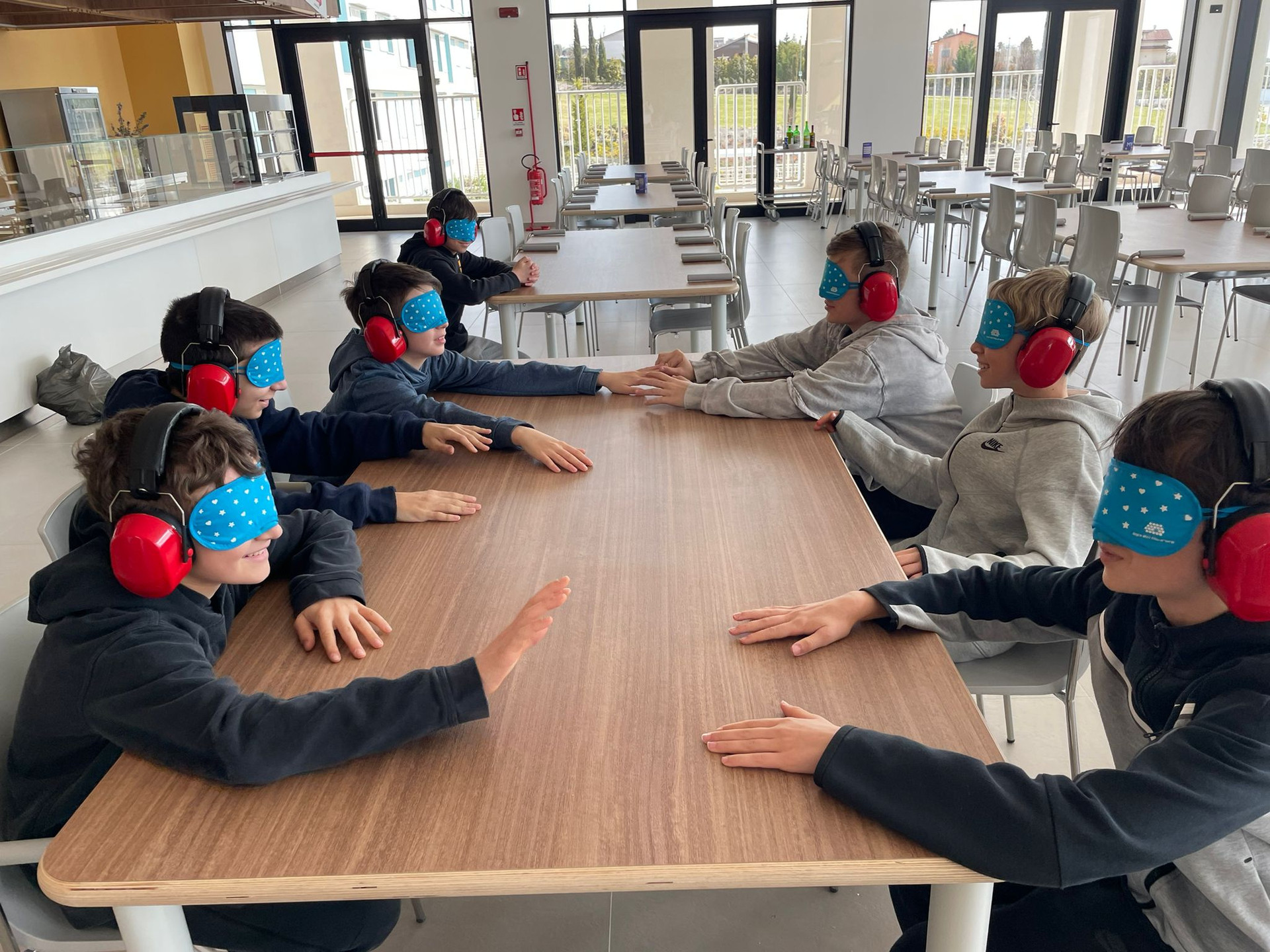 Gruppi e scuole in visita alla Lega del Filo d’Oro: i ragazzi sono seduti in un tavolo della mensa del Centro Nazionale di Osimo, indossano bende e cuffie, per un'esperienza nel buio e nel silenzio, provano a interagire tra loro