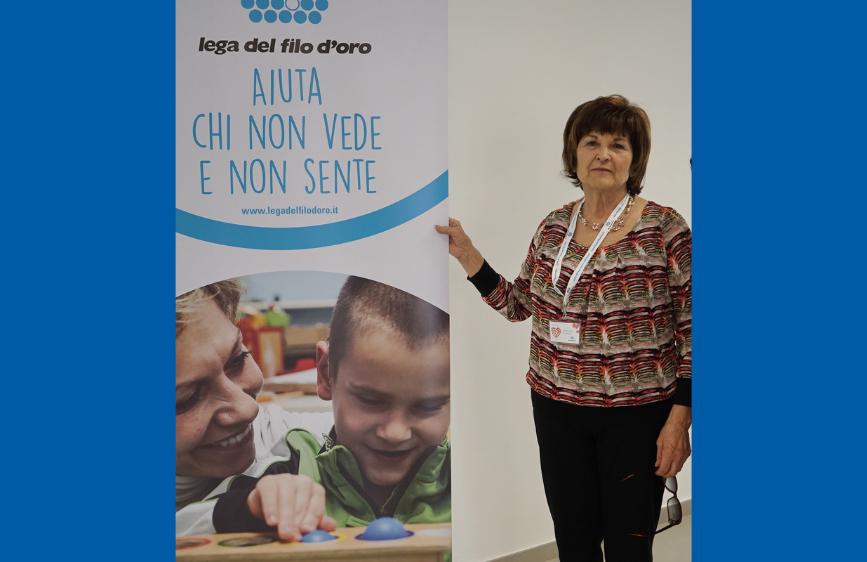 L'immagine mostra Liana Pulita, una sostenitrice, in piedi accanto a un banner verticale della "Lega del Filo d'Oro". Il banner, di colore blu, riporta il logo dell'organizzazione e lo slogan "AIUTA CHI NON VEDE E NON SENTE", con una foto in basso che ritrae un adulto che interagisce con un bambino.