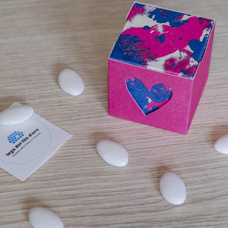 Bomboniera Cubo Pop composta da due scatoline in cartoncino pregiato inserite l'una nell'altra. Quella esterna è Fucsia, quella interna, visibile da un'apertura a forma di cuore, ha una fantasia bianca fucsia e blu ispirata ai disegni degli ospiti della Lega del Filo d'Oro