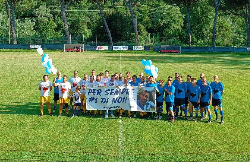 L'immagine mostra due squadre di calcio, una in maglia bianca e gialla e l'altra in maglia azzurra, schierate su un campo da gioco erboso, con alberi sullo sfondo. Al centro del gruppo, tengono uno striscione bianco con la scritta "PER SEMPRE #1 DI NOI!" e una foto di un uomo sorridente, in riferimento all'evento "Tutti per Anto". Alcuni palloncini bianchi e azzurri fluttuano sopra il gruppo, aggiungendo un tocco commemorativo all'atmosfera sportiva.
