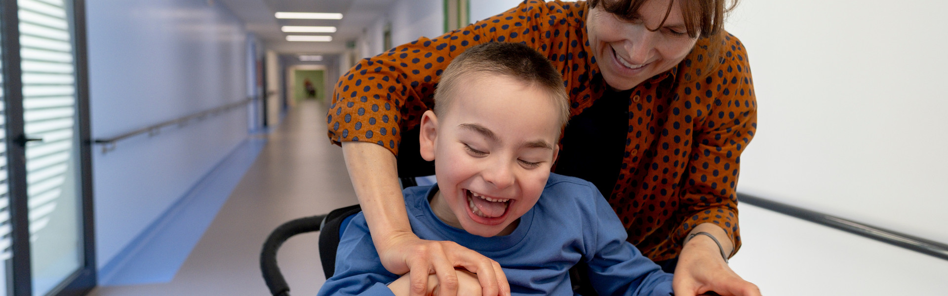 Edoardo, bambino utente seguito dalla Lega del Filo d'Oro, mentre viene portato a passeggio dall'educatrice per il Centro Nazionale di Osimo. Sorridono divertiti