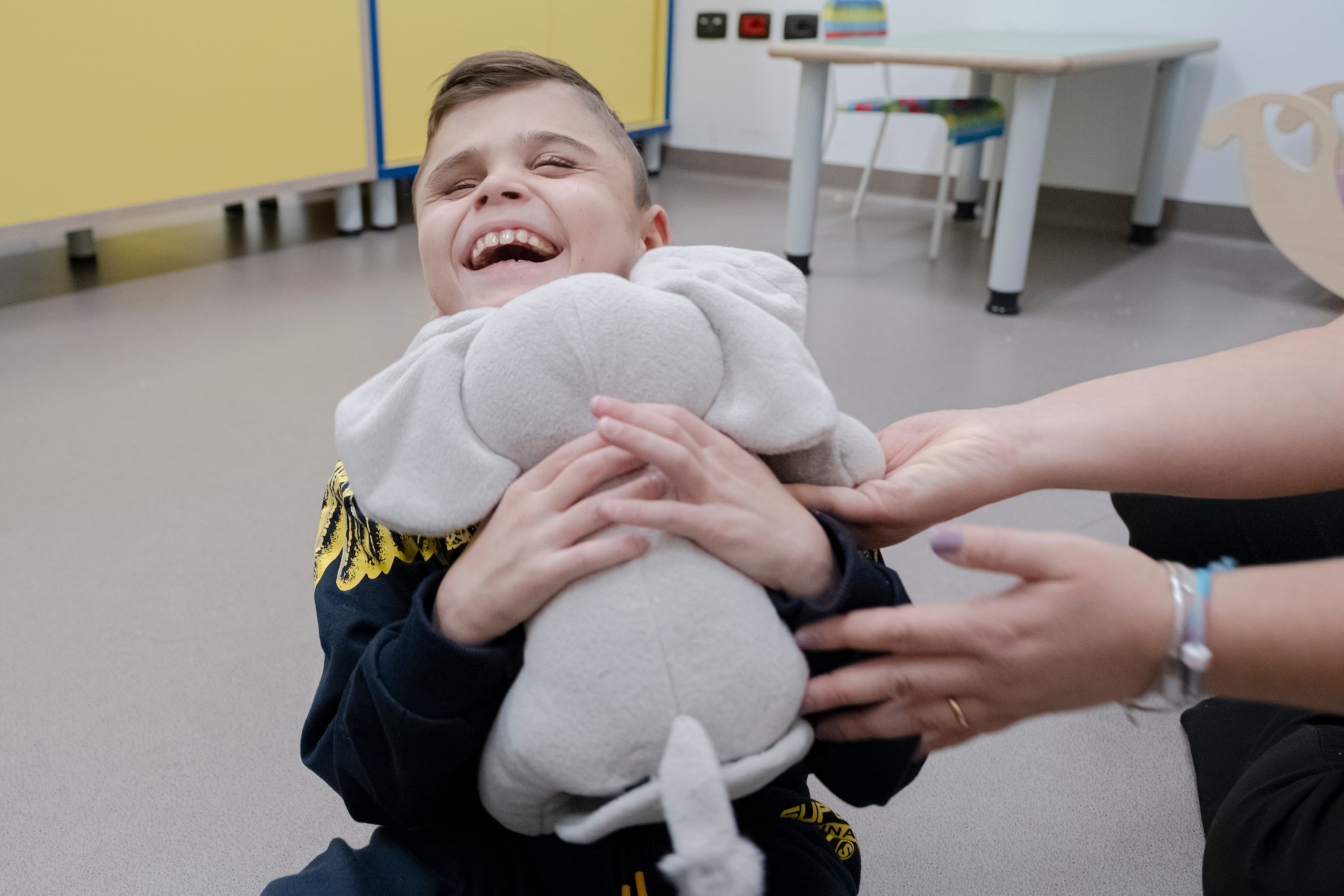 Matteo, bimbo seguito dalla Lega del Filo d'Oro, ride felice mentre abbraccia un peluche a forma di elefantino grigio.