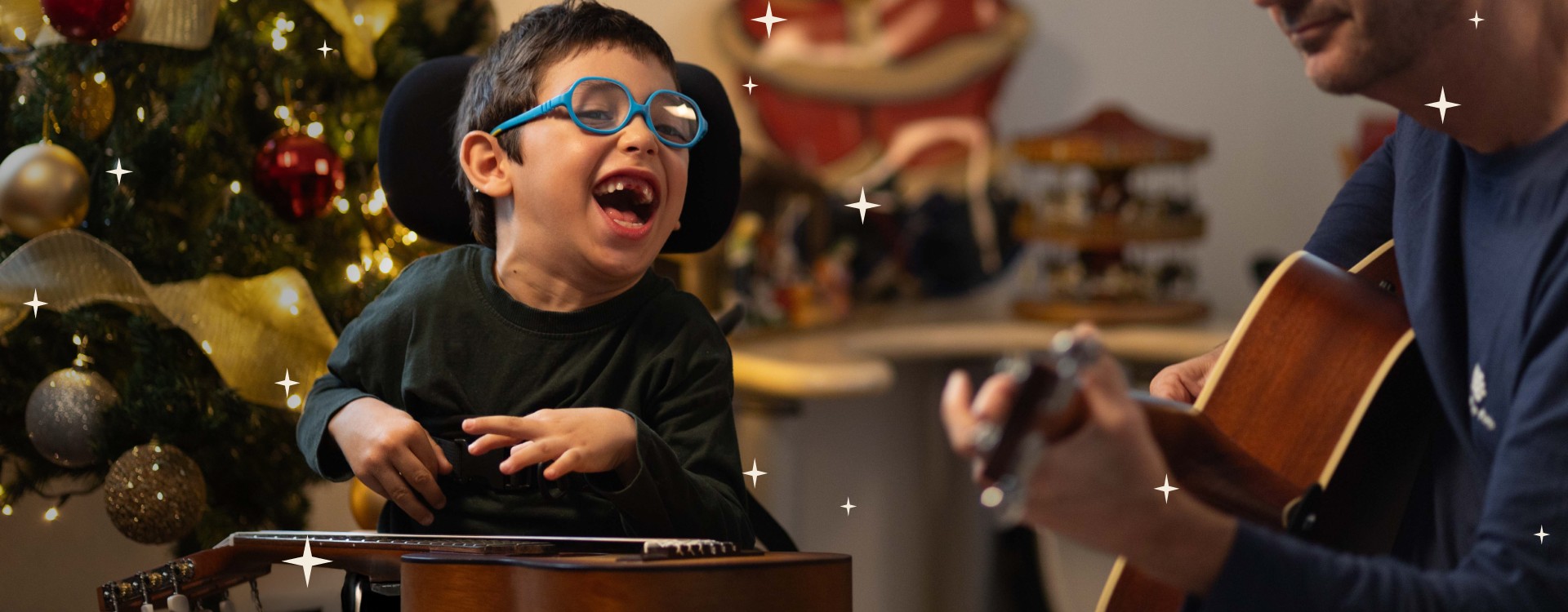 Bambino con disabilità motoria sorride accanto a un musicoterapista che suona la chitarra, con un albero di Natale sullo sfondo. Un momento di gioia