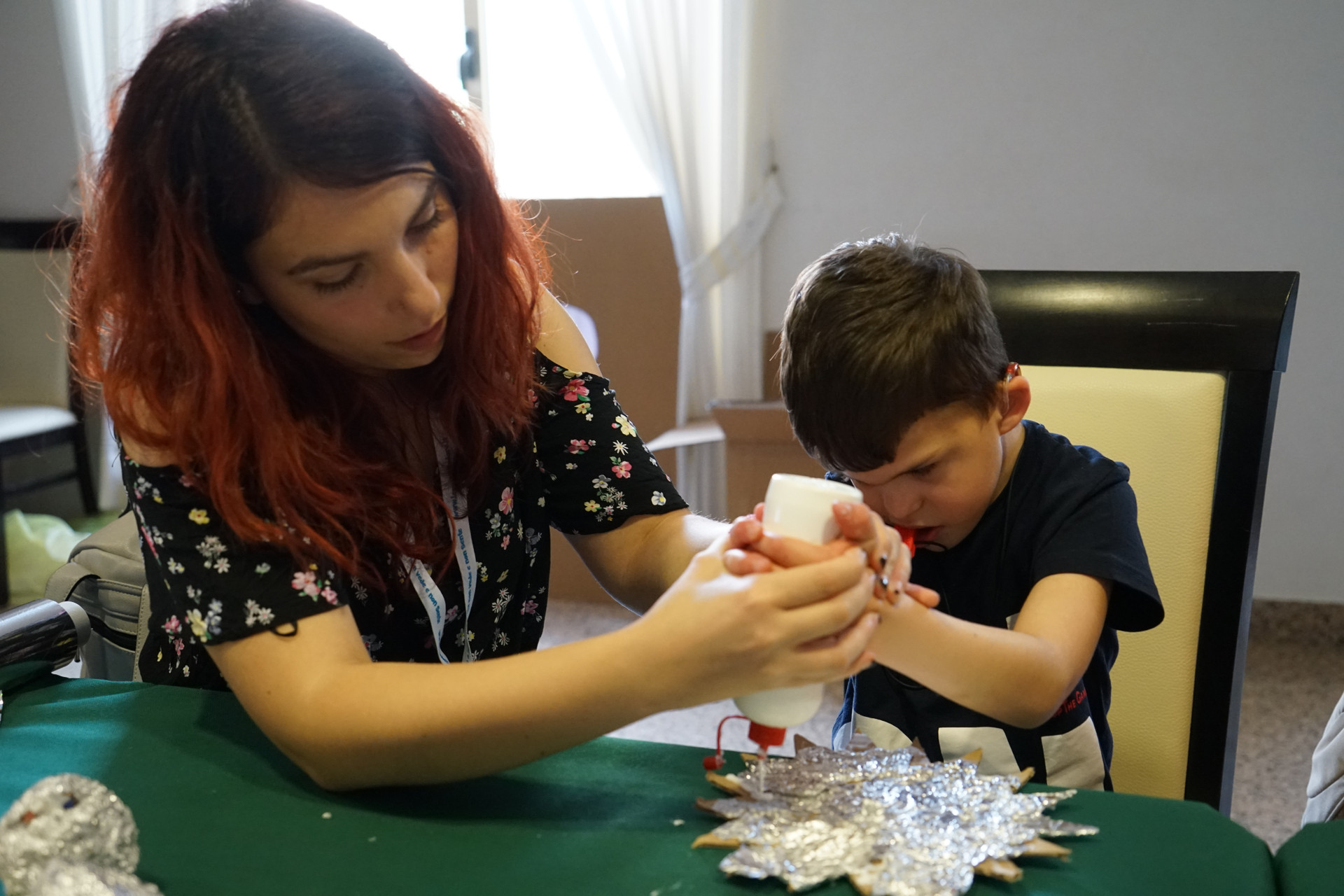 Giovane volontaria aiuta Agostino ad utilizzare la colla vinilica su un lavoretto di cartone e alluminio