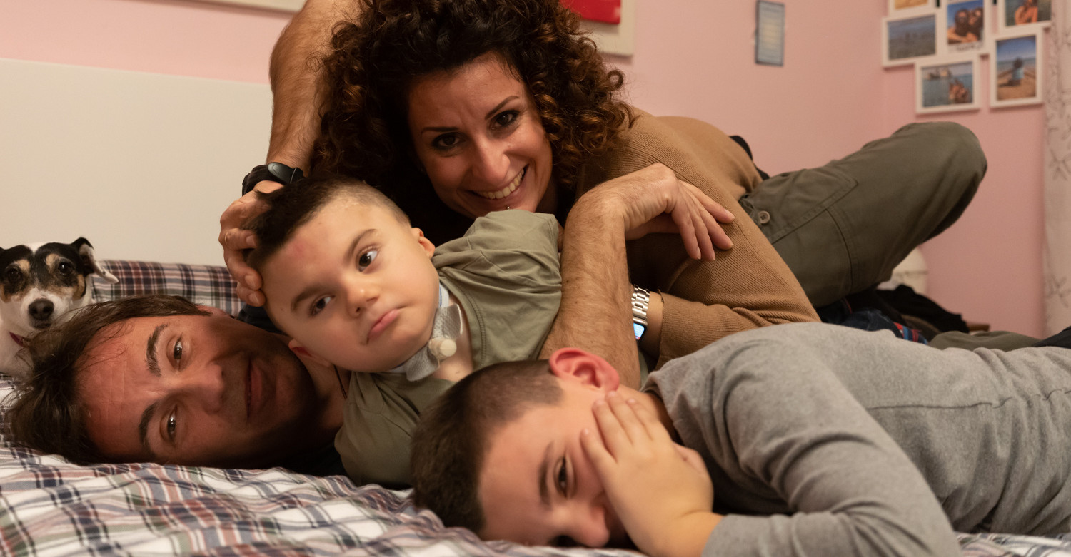 Foto della famiglia di Leonardo, un bambino affetto dalla sindrome di Charge, seguito dalla Lega del Filo d'Oro. Ci sono la mamma, il papà, Leonardo e il fratello maggiore e la loro piccola cagnolina. Sono stesi sul letto, sorridono alla camera