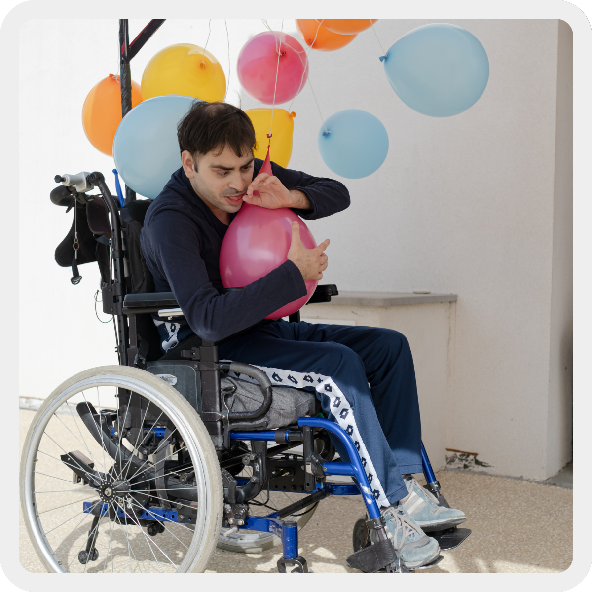 Simone, ragazzo con pluridisabilità di circa 25 anni, è sulla sua sedia a rotelle e abbraccia un palloncino rosa sorridendo. Intorno a lui altri palloncini colorati. 