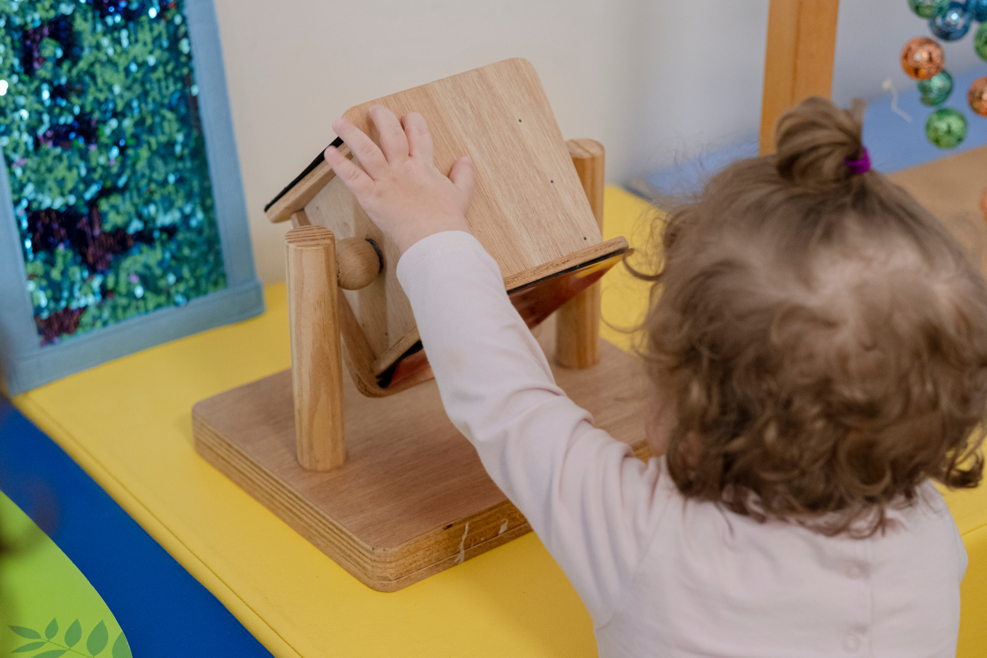 Una bambina bionda, vista di spalle, gioca con un gioco in legno. L'immagine ritrae un momento di gioco all’interno di un ambiente della Lega del Filo d’Oro.