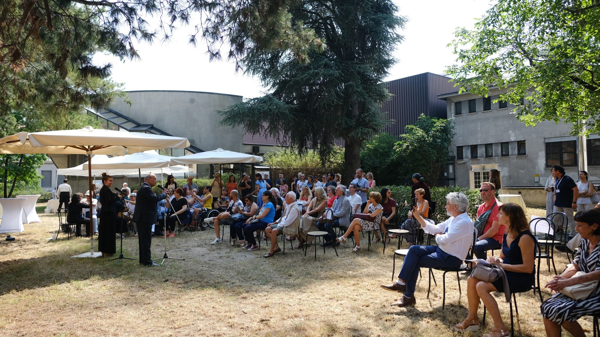Persone presenti all'inaugurazione della sede di Novara. Un gruppo di persone, sedute all'aperto, ascoltano il discorso del Presidente della Lega del Filo d'Oro.