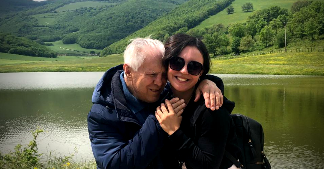 Tanya, volontaria della Lega del Filo d'Oro, abbraccia Angelo, utente sordocieco anziano, entrambi sorridono e si trovano in riva a un lago, immersi nel verde 