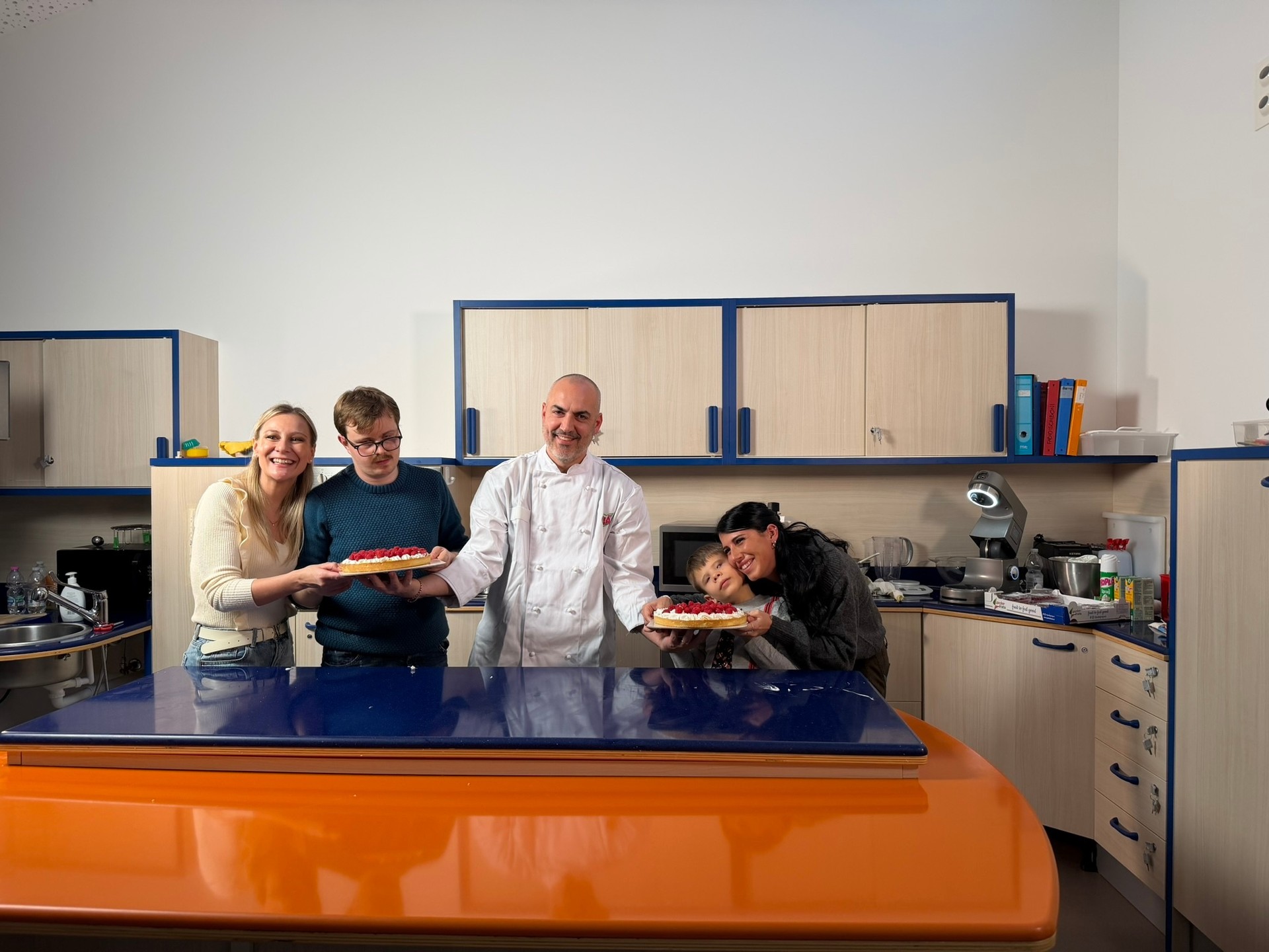 Nella cucina attrezzata del Centro NAzionale della Lega del Filo d'Oro, cinque persone sono dietro un piano di lavoro e mostrano duetorte con panna montata e fragole. il cuoco è al centro e indossa la divisa bianca, accanto a lui due ospiti del centro e due ragazze.
