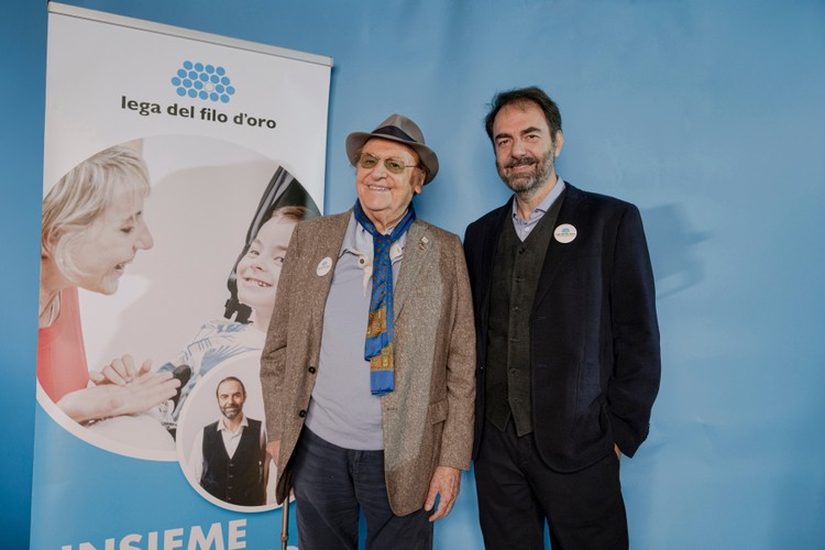Renzo Arbore e Neri Marcorè, testimonial della Fondazione LEga del Filo d'Oro, sono in piedi sorridenti fianco a fianco, vicino a un cartello verticale con logo e fotografie relative alla Fondazione