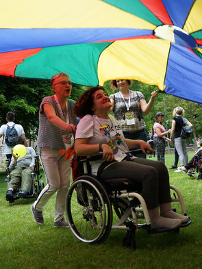 Una volontaria con una ragazza con pluriminorazione passa spingendo la carrozzina sotto un grande telo colorato tenuto sollevato da altri volontari