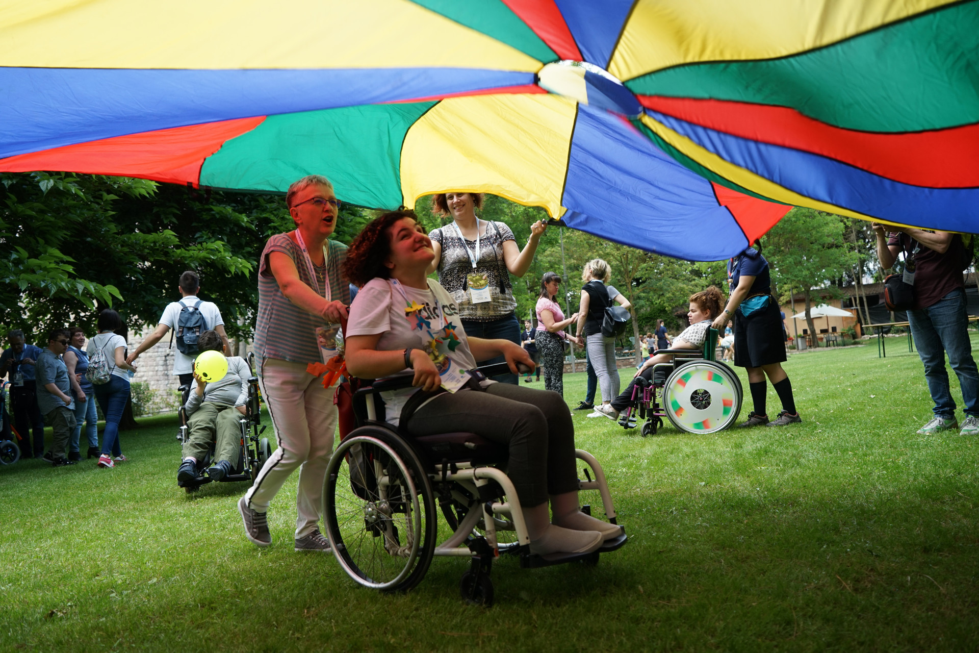 Una volontaria con una ragazza con pluriminorazione passa spingendo la carrozzina sotto un grande telo colorato tenuto sollevato da altri volontari