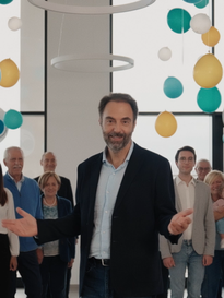 Foto che ritrae Neri Marcorè tra i palloncini, durante le riprese dello spot di Campagna del 60° anniversario della Lega del Filo d'Oro, nella mensa del Centro Nazionale di Osimo. Sullo sfondo molte persone che insieme sostengono la Fondazione, famiglie, utenti, personale specializzato, sostenitori