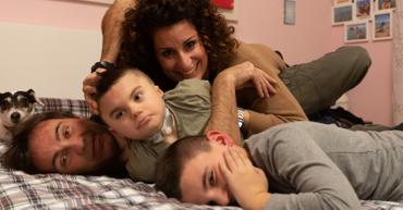 Leonardo, suo fratello, mamma, papà e il suo cagnolino giocano insieme sul lettone dei genitori e sorridono felici per una foto di gruppo.