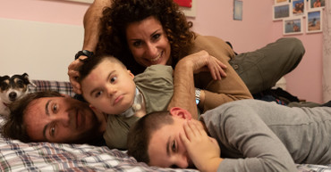 Leonardo, suo fratello, mamma, papà e il suo cagnolino giocano insieme sul lettone dei genitori e sorridono felici per una foto di gruppo.
