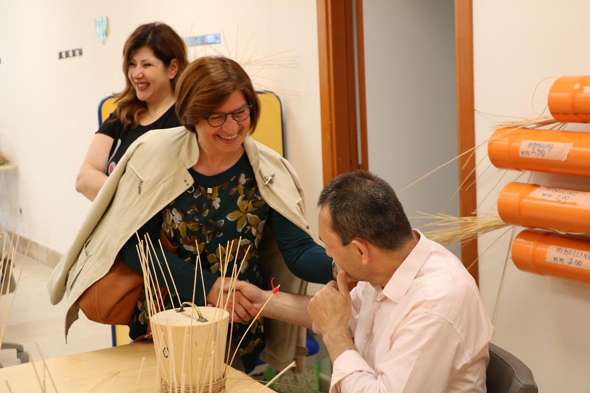 Giornata del Sostenitore - Incontro utente con sostenitrice - L'utente sordocieco adulto interrompe il suo lavoro con il midollino per stringere la mano alla sostenitrice sorridente