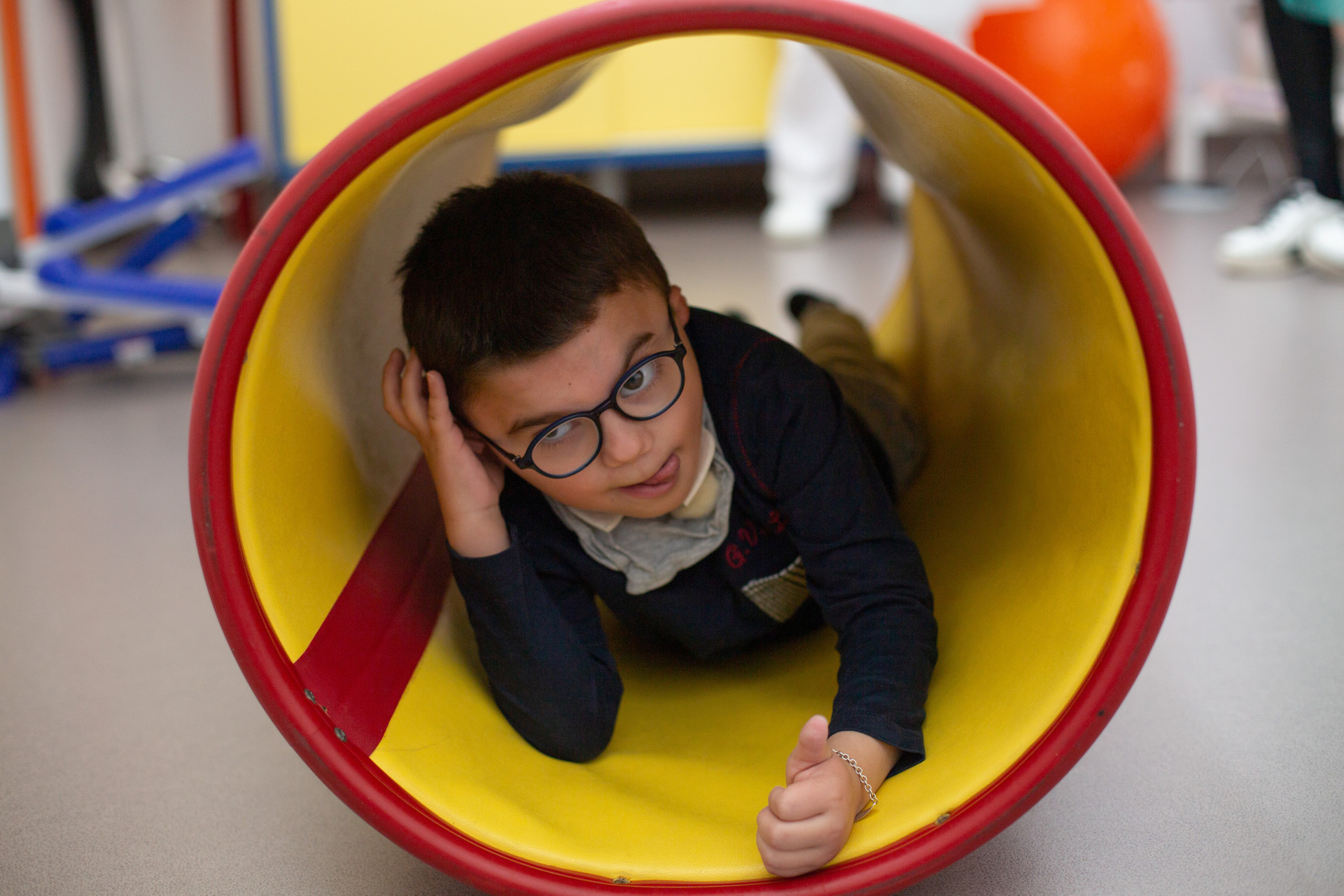 Leonardo bambino di 5 anni si affaccia da dentro un grande tubo rosso e giallo, durante una seduta di fisioterapia