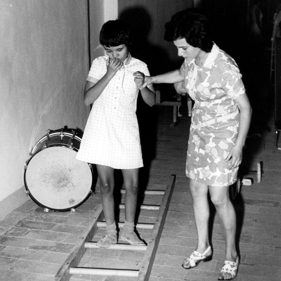 Una bambina sordocieca viene aiutata da un'operatrice in un esercizio. C'è una scala appoggiata a terra e lei deve camminare senza toccare i pioli. Foto degli anni '70 e in bianco e nero.