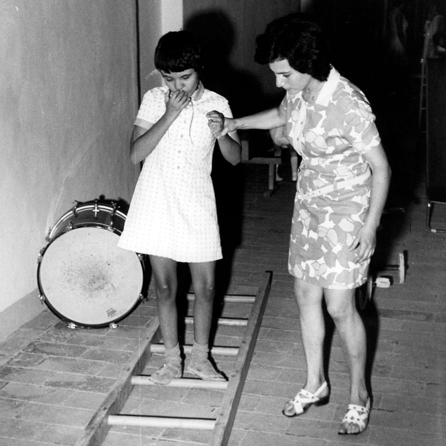 Una bambina sordocieca viene aiutata da un'operatrice in un esercizio. C'è una scala appoggiata a terra e lei deve camminare senza toccare i pioli. Foto degli anni '70 e in bianco e nero.