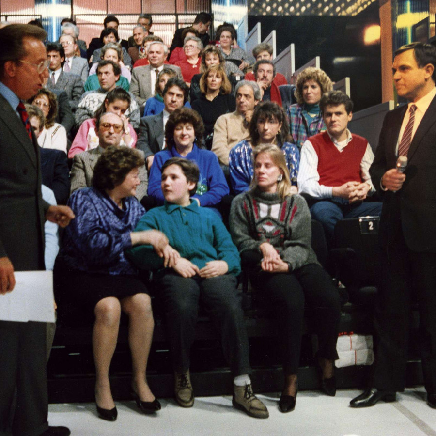Alcuni utenti sordociechi sono nello studio della trasmissione Penthatlon di Mike Bongiorno. Davanti a loro il presidentatore, in piedi, ascolta un altro uomo, anche lui in piedi e col microfono in mano.