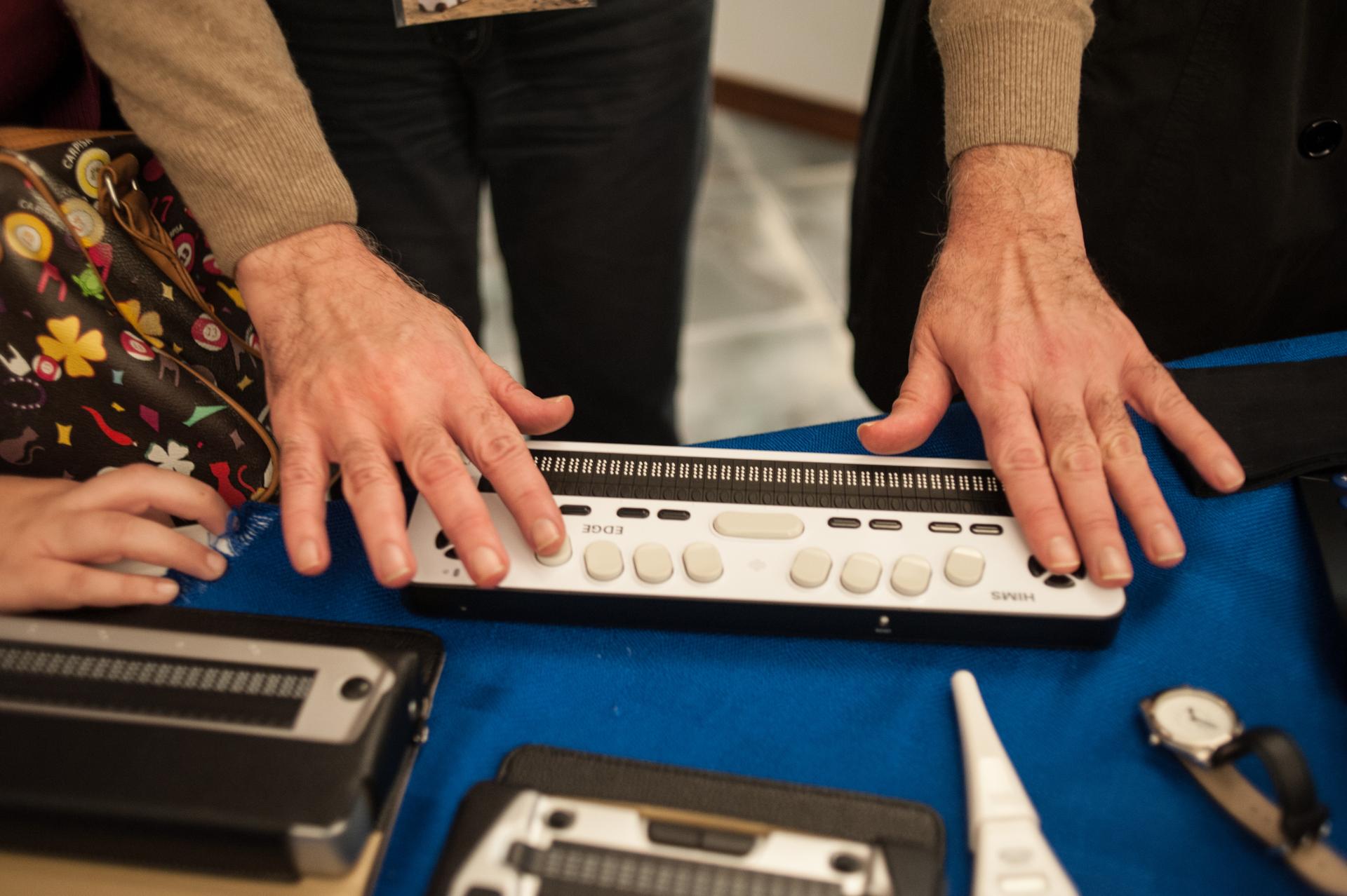 Dettaglio ravvicinato di mani maschili che scrivono su una barra braille. La barra braille è poggiata su un tavolo.