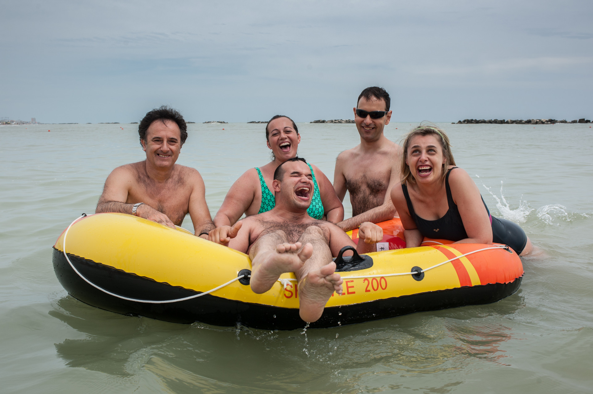 Soggiotni estivi al mare: un utente ride di gusto galleggiando su un gommone mentre volontari ed altri utenti, appoggiati intorno al gommone, ridono insieme a lui