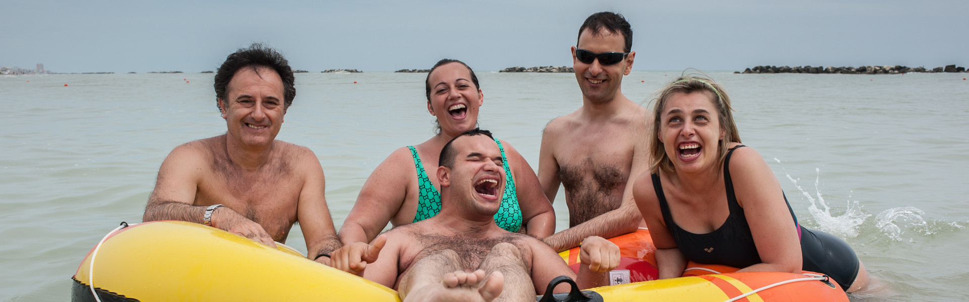 Soggiotni estivi al mare: un utente ride di gusto galleggiando su un gommone mentre volontari ed altri utenti, appoggiati intorno al gommone, ridono insieme a lui