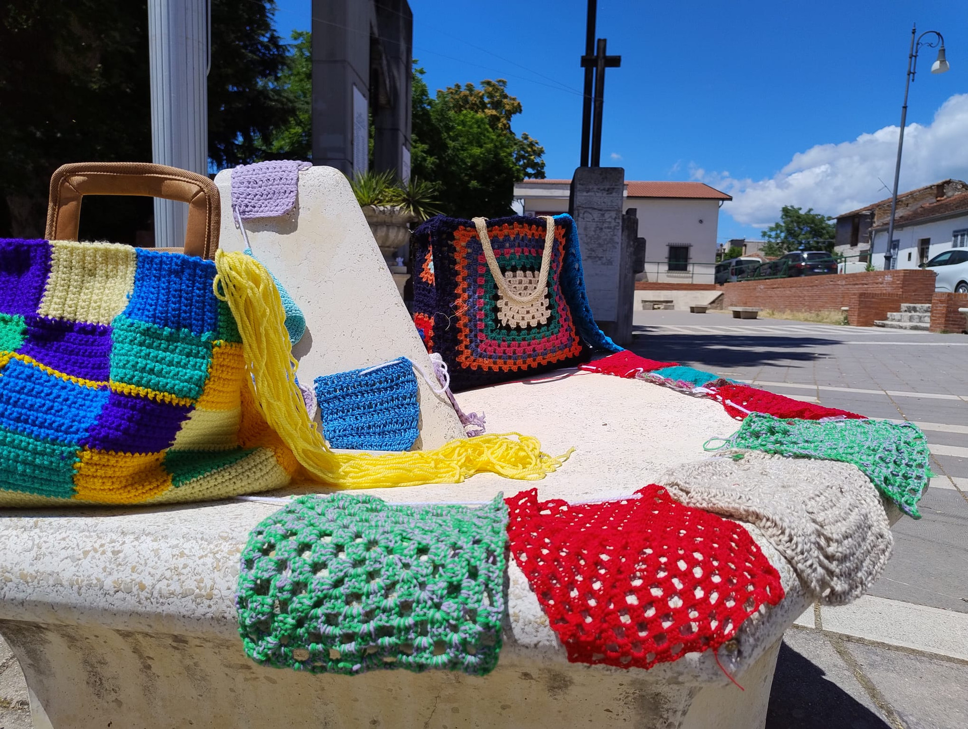 Dettaglio di una panchina decorata con creazioni a maglia per lo Yarn Bombing promosso dalla Lega del Filo d’Oro. Presenti mattonelle colorate: verdi, rosse, bianche, celesti, blu e lilla e due borse multicolor fatte all’uncinetto.