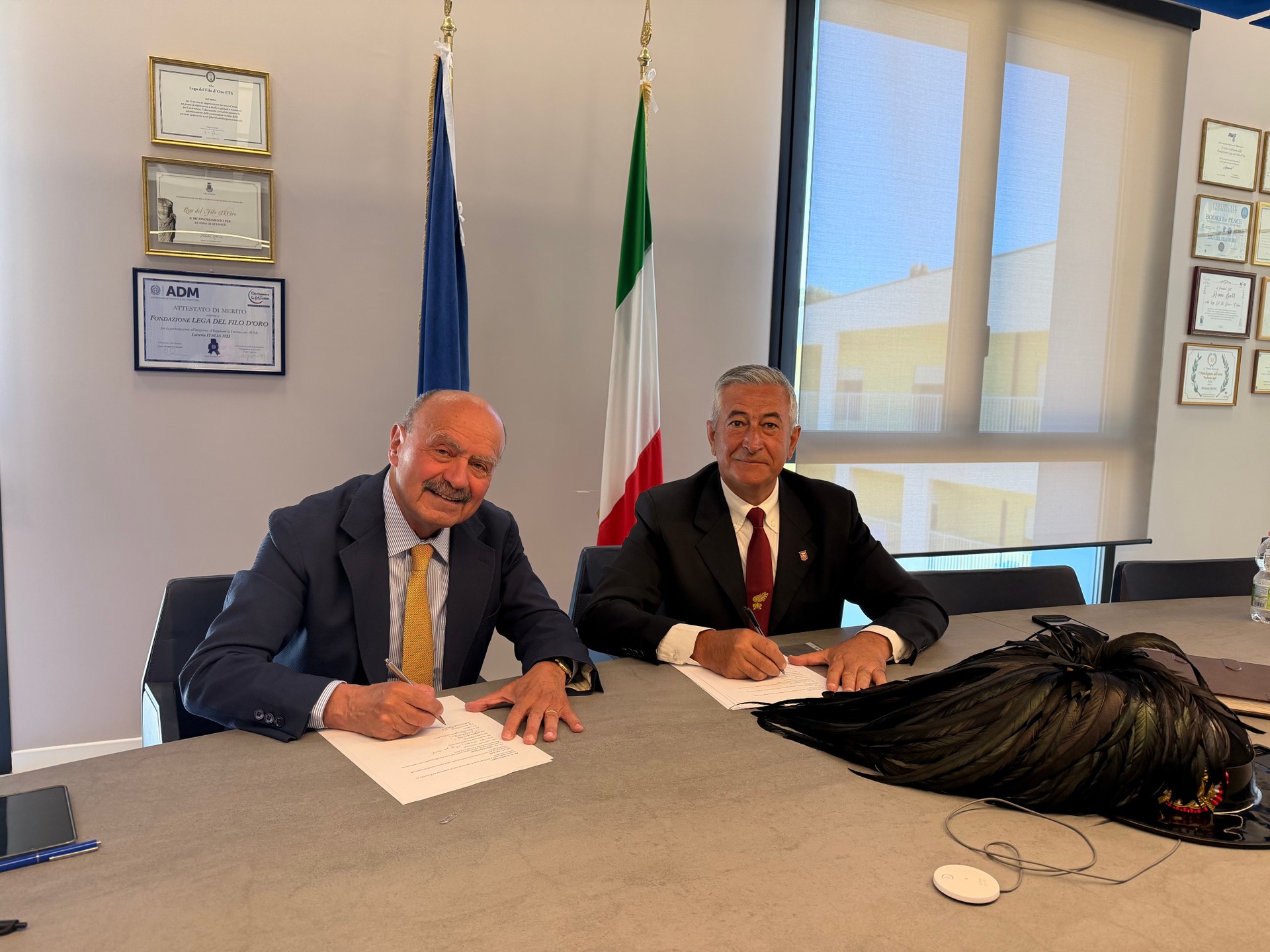 Il Presidente della Lega del Filo d'Oro (a sinistra) e Presidente dell’Associazione Nazionale Bersaglieri (a destra), siedono a un tavolo mentre firmano un Protocollo di Intesa finalizzato a promuovere, a livello nazionale e territoriale, la promozione e realizzazione di iniziative sociali, culturali, educative e solidali. Sullo sfondo si vedono la bandiera italiana e certificati incorniciati. Sul tavolo il cappello piumato tipico dei bersaglieri