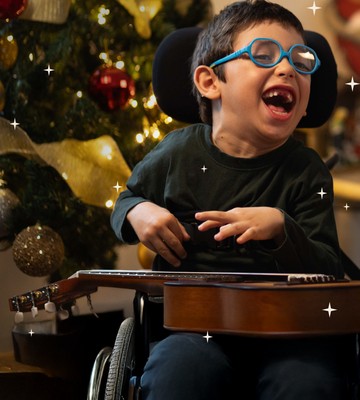 Bambino con disabilità motoria sorride accanto a un musicoterapista che suona la chitarra, con un albero di Natale sullo sfondo. Un momento di gioia