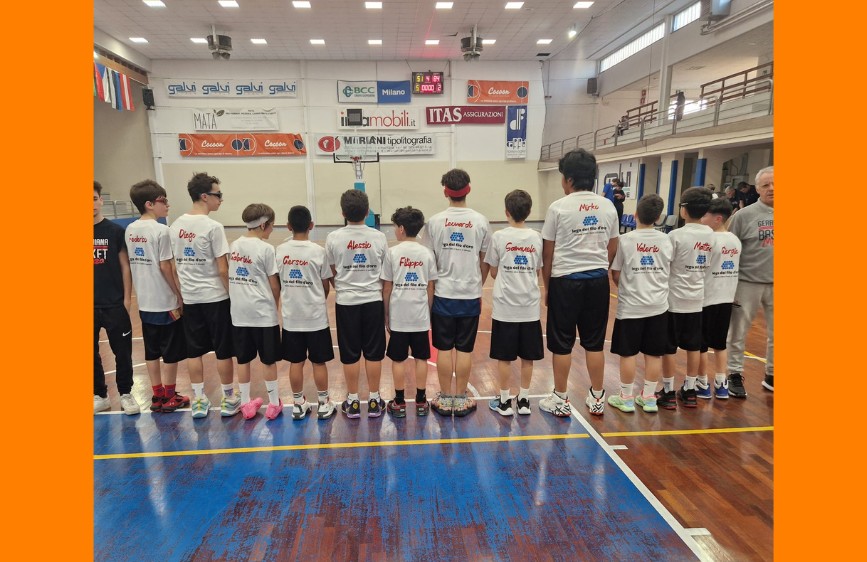 Squadra giovanile in palestra vista di spalle, con magliette bianche personalizzate con i nomi dei ragazzi. Sul parquet di un campo da basket, durante un evento sportivo.