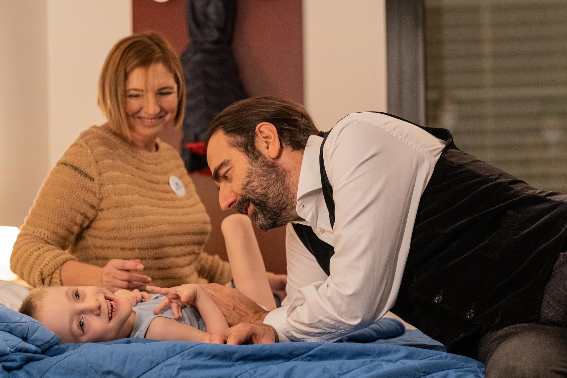 Gabriele, bambino ospite della Lega del Filo d'Oro, Neri Marcorè e un'operatrice posano sorridenti. L'operatrice è seduta sul letto dove Gabriele è disteso supino e ride, mentre Neri lo guarda e accarezza con affetto.